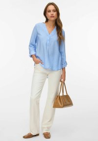 Femme portant un chemisier à rayures bleu clair, un pantalon blanc et des chaussures marron, tenant un sac à main marron dans la main gauche.