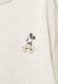 Sudadera de color beige claro con un gráfico en negro y amarillo de Mickey Mouse, sonriendo y corriendo, con una textura suave y un corte relajado.