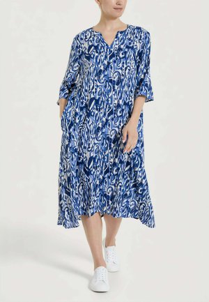 Donna che indossa un vestito largo fantasia blu e bianco al ginocchio, con maniche a tre quarti e sneakers bianche, in piedi con le mani nelle tasche.