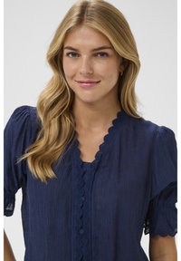 Usmievavá žena s dlhými blond vlasmi, oblečená v tmavomodrej textúrovanej blúzke s krajkovým lemom so zubatým okrajom a nafúknutými krátkymi rukávmi.