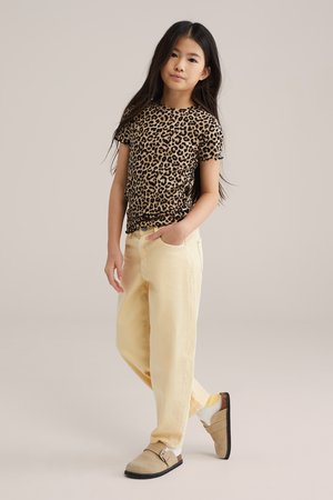 Mladé dievča stojí s jednou rukou v vrecku, oblečené v krátkosúložnej košeli s leopardím vzorom, svetložltých nohaviciach, bielych ponožkách a béžových slip-on topánkach.