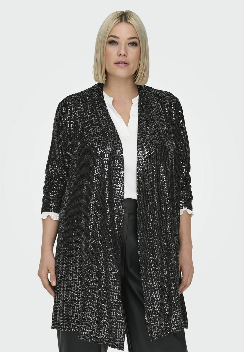 Must mustriga pikk jakk avatud ees, kolmeveerandvarrukate ja tekstureeritud mustriga, kantud valge bluusiga ja mustade pükstega.