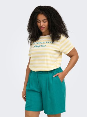 Femme aux cheveux bouclés portant un t-shirt rayé jaune et blanc avec un texte et un short taille haute couleur sarcelle, debout avec une main dans la poche.