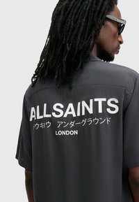Grijs shirt met korte mouwen en een zachte structuur, voorzien van "ALLSAINTS" en Japanse tekst in het wit gedrukt op de achterkant.