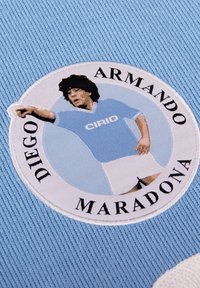 Tela azul clara con textura acanalada que presenta un parche bordado circular. El parche muestra a un jugador de fútbol con una camiseta azul clara con el texto "CIRIO".
