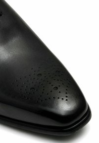 Zwarte leren dresschoenen met een spitse neus, voorzien van decoratieve perforaties in een wervelend patroon op de bovenkant. Gladde textuur en subtiele glans.