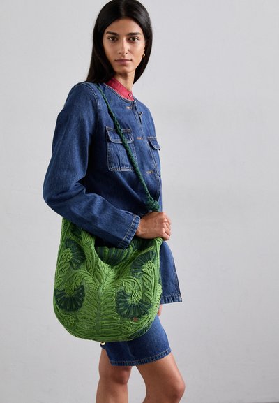 Geantă de umăr tricotată verde, cu modele florale intricate și curea împletită, expusă pe un fundal neutru. Un outfit din denim completează designul.
