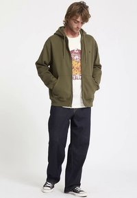 Olive green zip-up hoodie with a front pocket, worn over a graphic t-shirt. Paired with dark blue straight-leg jeans and black sneakers.