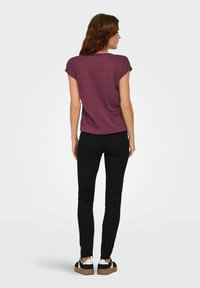 Une femme aux longs cheveux bruns se tient de dos, portant un haut bordeaux à manches courtes, un jean skinny noir et des baskets noir et blanc.