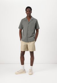 Camiseta de manga corta de color gris texturizado con un bolsillo en el pecho, combinada con pantalones cortos beige. El modelo lleva zapatillas beige y calcetines blancos.