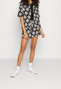 Kobieta w czarnej kwiatowej koszulce z krótkim rękawem i dopasowanych szortach, z czarnym topem pod spodem, w białych skarpetkach i białych trampkach, stojąca przy białej ścianie.