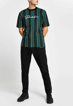 Mann steht, trägt schwarze Hose, schwarze Sneakers und ein grün-schwarzes vertikal gestreiftes kurzärmeliges Hemd mit weißem Script-Text.