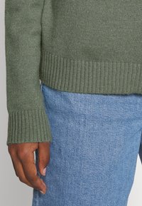 Olivgrön stickad tröja med ribbade ärmar och fåll, kombinerad med ljusblå jeans. Visar närbild av ärmen och texturdetaljer.