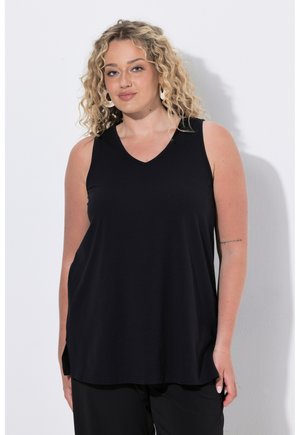 Femme blonde aux cheveux bouclés portant de grandes boucles d’oreilles créoles, un haut noir sans manches et un pantalon noir, debout contre un mur blanc.