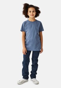 Blå bomuld t-shirt med korte ærmer, kombineret med mørkeblå denimjeans og hvide lærredssneakers, stående på en neutral baggrund.