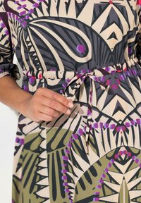 Blumenmuster-Kleid aus leichtem Stoff mit einer taillierten Taille und einer Schleife, sowie langen Ärmeln. Die Farben umfassen Lila, Grün und Beige.