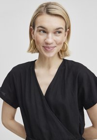 Robe noire enveloppante à manches courtes et décolleté en V profond. Fabriquée dans un tissu léger avec une texture subtile, elle est dotée d'une ceinture à nouer.