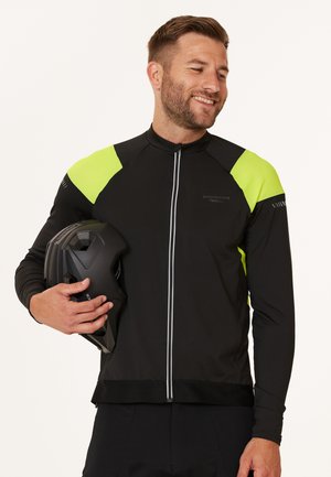 Schwarze Fahrradjacke mit leuchtend gelben Netz-Accents, voller Reißverschluss, reflektierenden Streifen und Kragen-Details. Modell hält einen schwarzen Helm.