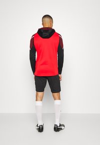 Rotes Sportshirt mit schwarzen langen Ärmeln, schwarzen Shorts, weißen Socken und schwarzen Fußballschuhen, ausgestattet mit einer Kapuze und strukturierten Schulterakzenten.