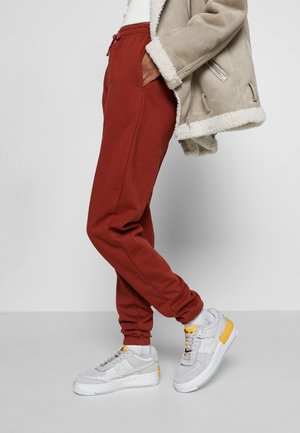 Pantalon de jogging rouge avec poches latérales, bas resserrés et taille ajustable, associé à des baskets grises avec des accents jaunes.