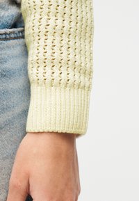 Κοντινή λήψη ενός χεριού που φοράει μανίκι από εκρού πλεκτό πουλόβερ δίπλα σε ανοιχτό μπλε τζιν τζιν παντελόνι πάνω σε λευκό φόντο.