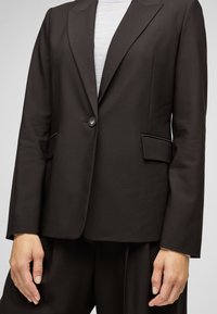 Veste noire cintrée avec une fermeture à un seul bouton, des revers crantés et deux poches avant, fabriquée en tissu structuré à texture lisse.