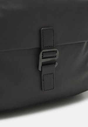 Gros plan sur le rabat d'un sac noir maintenu par une sangle noire avec des bords cousus et une boucle en métal noir mat.