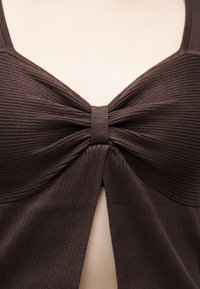 Κοντινό πλάνο καφέ ριμπ πλεκτού τοπ με στριφτό κόμπο και λεπτομέρεια με ανοιχτή τρύπα στο κέντρο μπροστά.