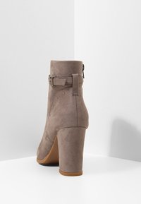 Kotníková bota z taupe semiše s vysokým blokovým podpatkem, dekorativním bočním prošitím, zapínáním na zip a nastavitelným páskem s přezkou kolem kotníku.