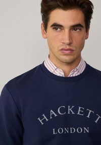 Marine sweatshirt med rund hals og grå tekst "HACKETT LONDON." Båret over en pink- og hvidstribet skjorte. Glat tekstur.