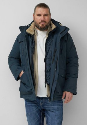 Brodaty mężczyzna ubrany w wielowarstwową zimową odzież: niebieska parka z kapturem wyłożonym futrem, beżowy sweter na zamek, biała koszulka i niebieskie dżinsy.