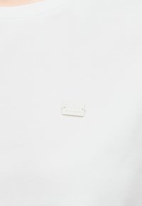 Valge puuvillane T-särk ümarate kaelustega, rinnataskul väike hõbedane silt, millel on lõike ja brändi detailid. Sile tekstuur.