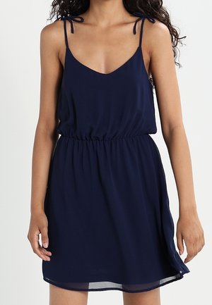 Femme portant une robe bleu marine sans manches avec des fines bretelles et une taille élastique, debout devant un fond clair uni.