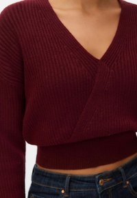 Gros plan sur une personne portant un pull cache-cœur côtelé bordeaux foncé avec un col en V et un jean taille haute bleu foncé.