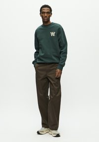Dark green sweatshirt with beige "W" logo, paired with brown trousers. Model wears light gray and black sneakers. Simple, casual ensemble.