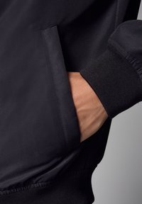 Dettaglio di una giacca bomber nera che mostra una mano in una tasca laterale, polsini a costine neri e una texture di tessuto liscia e opaca.