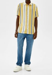 Camisa de botones de manga corta en amarillo con rayas verticales blancas y azul marino; combinada con jeans azul y zapatillas blancas. Textura de tela suave.