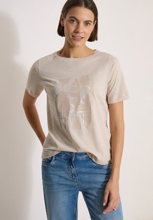 Femme portant un t-shirt graphique beige et un jean bleu, debout avec une main dans la poche devant un fond clair uni.
