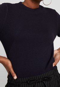 Ciemnogranatowa krótka dzianinowa bluzka z krótkim rękawem o prążkowanej fakturze i okrągłym dekolcie. Na ramionach znajdują się ukośne prążkowane panele.