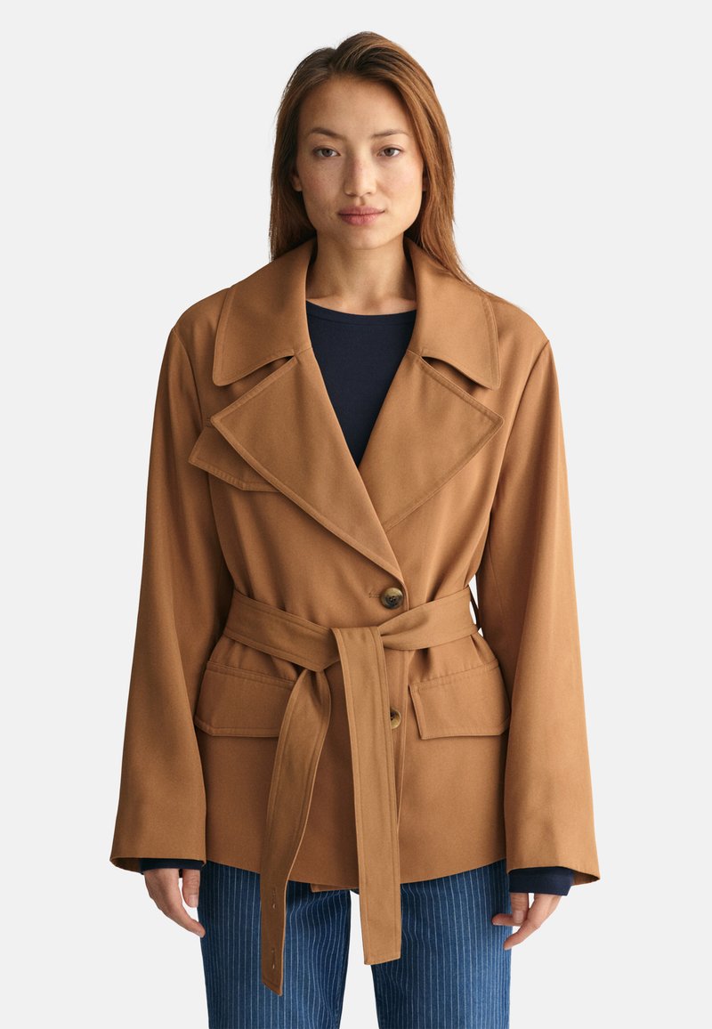 Manteau marron ceinturé avec un large col, fermeture par boutons et deux poches avant. Possède un design structuré et un tissu doux. Pantalons à rayures horizontales.