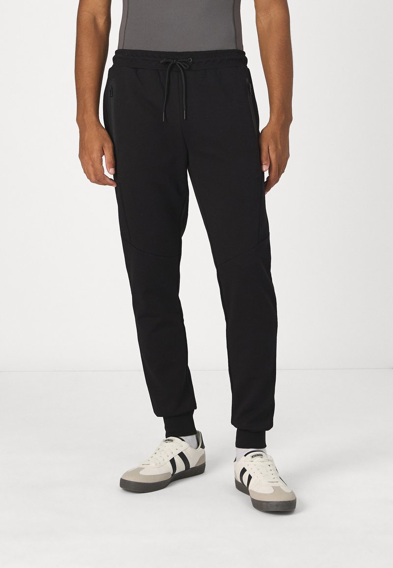 Black jogger pants made of soft fabric, featuring a drawstring waistband and zippered side pockets, paired with cream and gray sneakers.