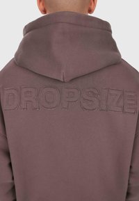 Egy személy hátulról, aki barna kapucnis pulóvert visel, amelyen nagy, hímzett "DROPSIZE" felirat található a háta felső részén, a kapucni alatt.