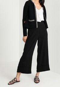 Cardigan nero corto con rifiniture argentate, indossato sopra una blusa bianca con volant, abbinato a pantaloni neri a gamba larga e sandali neri con cinturini.