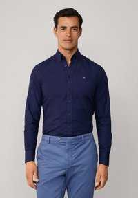 Donkerblauwe overhemd met lange mouwen en een kraag, voorzien van een klein geborduurd logo. Gecombineerd met lichtblauwe broek.