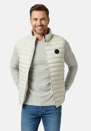 Lachende man die een lichtgrijs gewatteerd vest draagt over een grijs shirt met lange mouwen en een blauwe spijkerbroek, met zijn handen achter zijn rug.