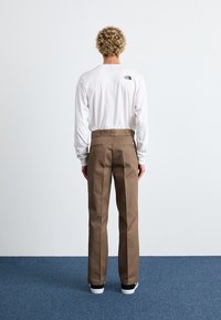 Brown pants with a straight fit, made of sturdy fabric, featuring two back pockets and a zipper detail. Paired with a white long-sleeve shirt.
