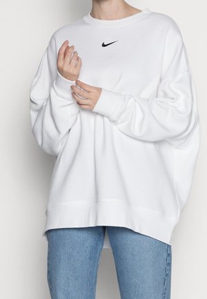 Personne portant un sweat-shirt Nike blanc ample et un jean bleu, ajustant la manche avec les deux mains visibles.