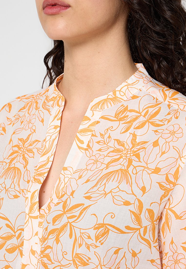 Biała bluzka z pomarańczowym wzorem w kwiaty, kołnierzyk mandarynki i listwą guzikową. Tkanina wydaje się lekka o gładkiej fakturze.