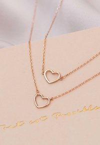 Gouden ketting met twee verbonden hartbedels aan delicate kettingen, liggend op een beige achtergrond met gouden letters.