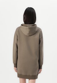 Rochie din bumbac maro, cu maneci lungi, glugă spațioasă și manșete nervurate. Textură netedă, se oprește deasupra coapsei, cu o croială lejeră.